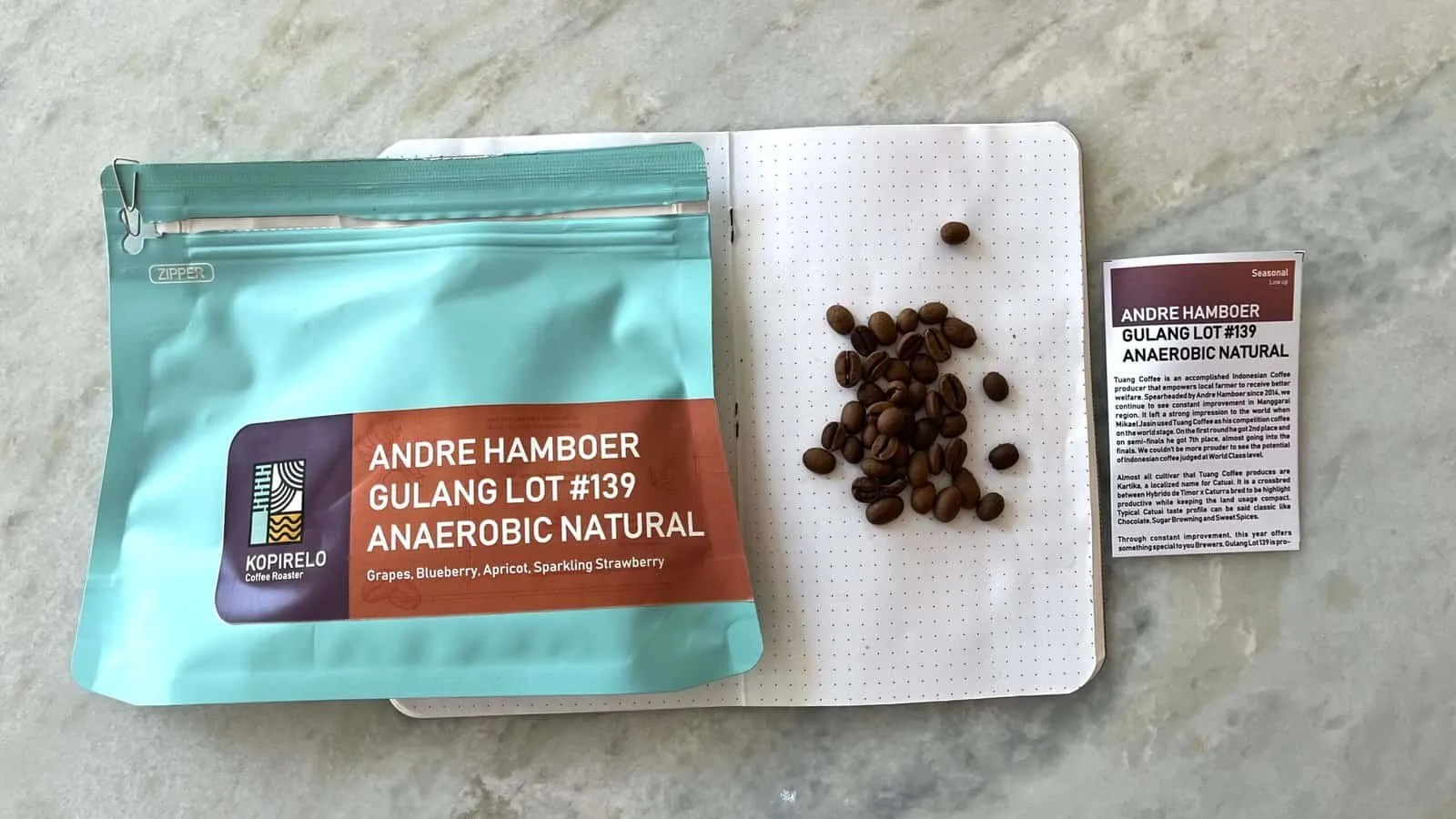 Alt text: Raw coffee beans displayed on a white notebook alongside a bag of Andre Hamber Gulang Lot #139 Anaerobic Natural coffee with detailed description.