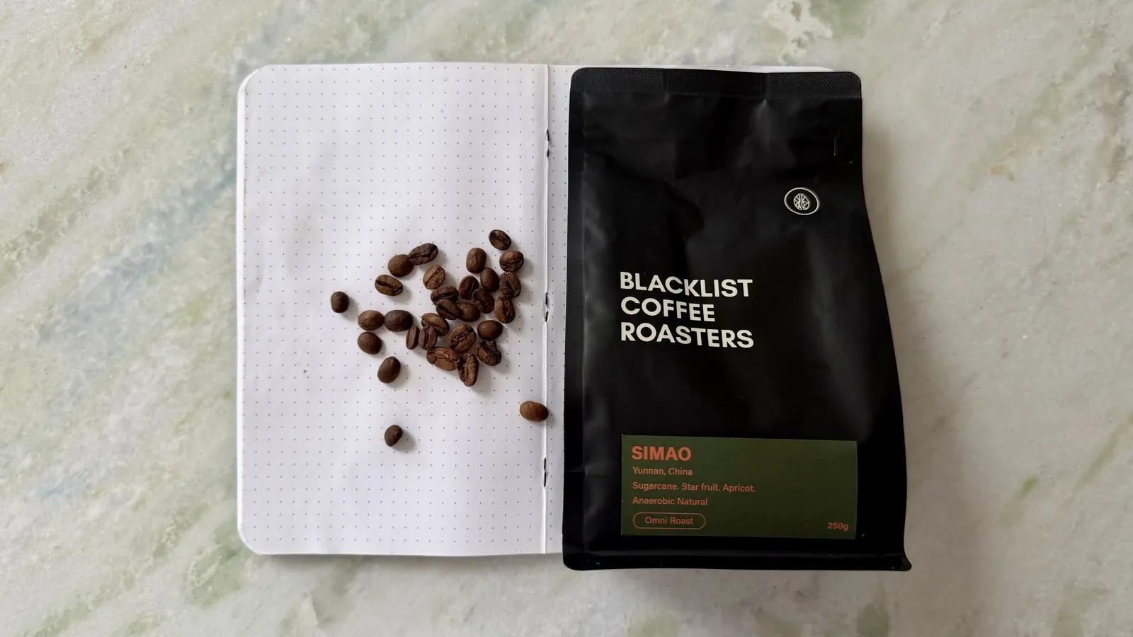 Close-up of roasted coffee beans on an open notebook with a black coffee bag from Blacklist Coffee Roasters in the background, showcasing high-quality specialty coffee.