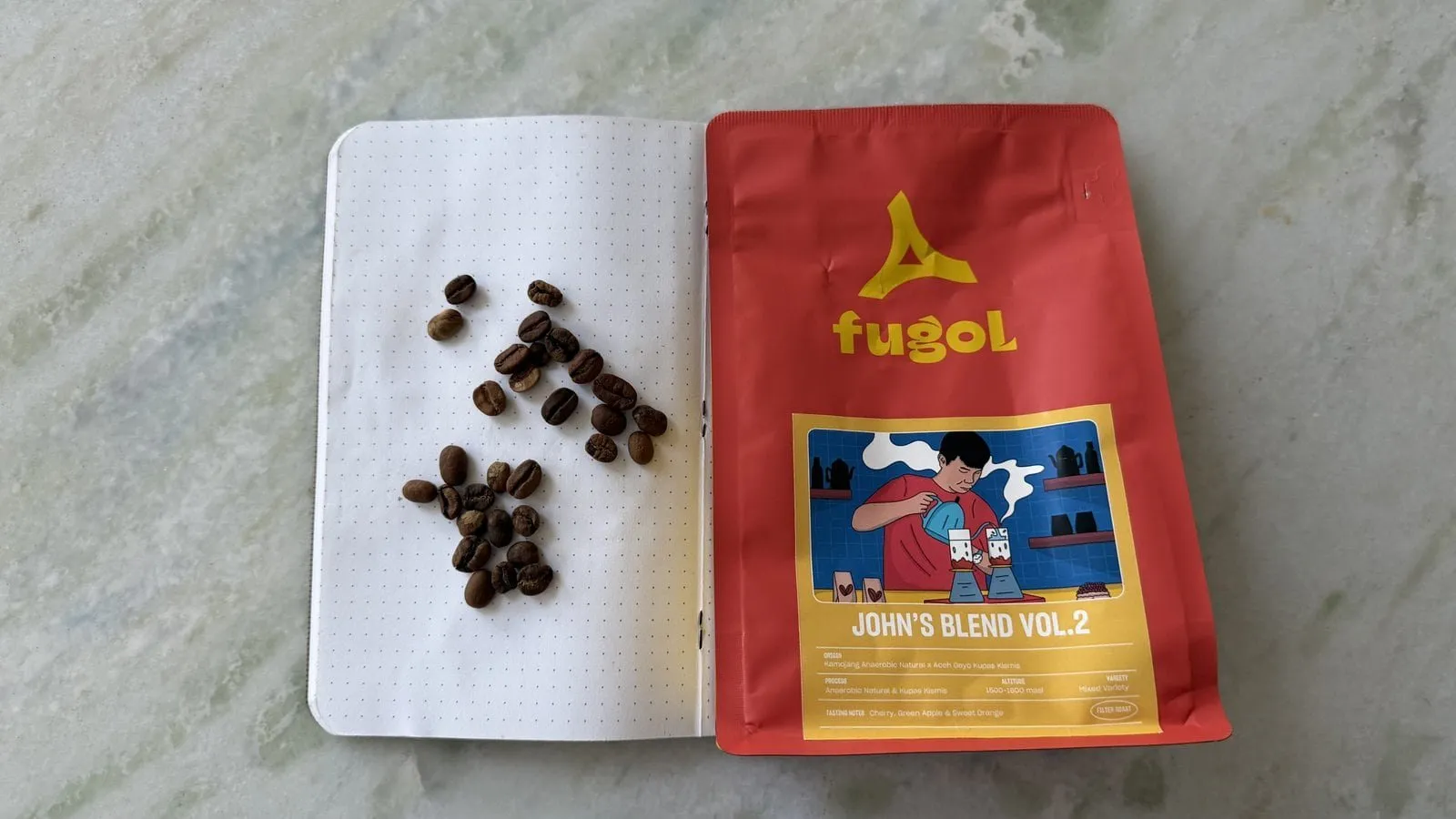 Coffee beans arranged on a dotted notebook page next to a red FUGOL coffee bag featuring a colourful illustration.