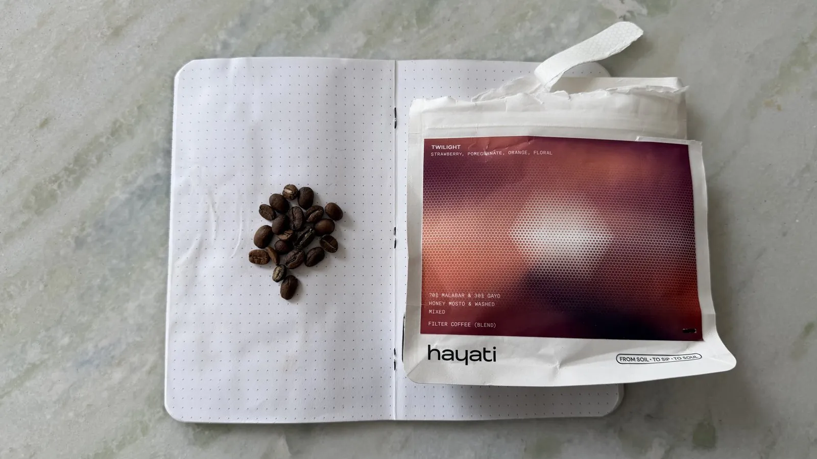 Close-up of coffee beans on an open notebook with a coffee package on a light grey surface.