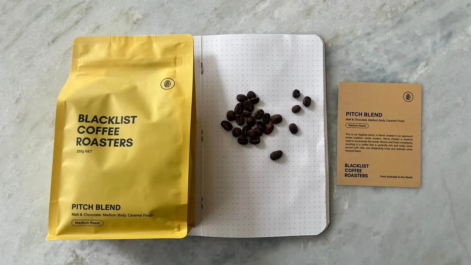 Alt text: Yellow coffee bag labeled "BlackList Coffee Roasters" with coffee beans and a tasting note card on a marble surface.